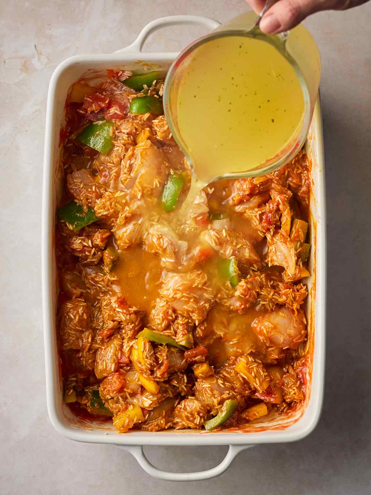 An oven dish with chicken, rice and curry paste, with a hand pouring over chicken stock from a jug.