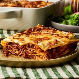 A side aspect of a plate with a slice of lasagne on, with the rest of the dish in the background.