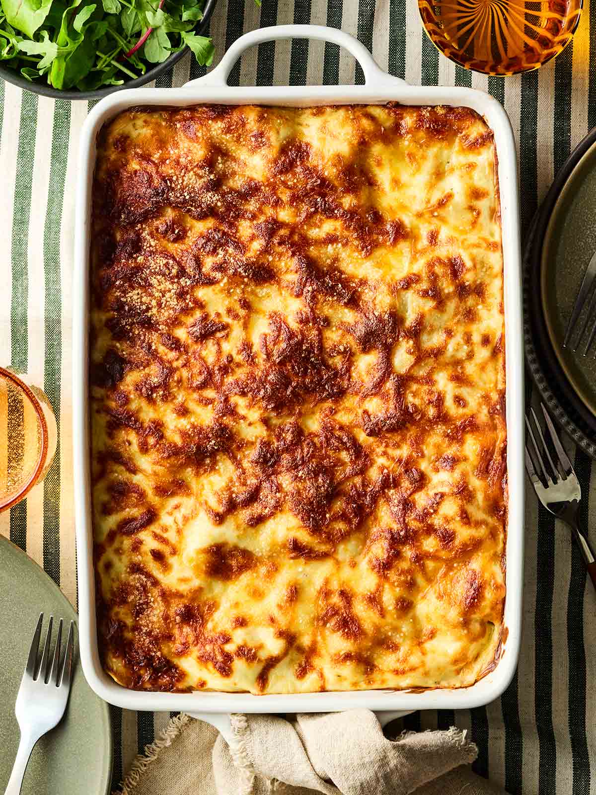 A bird's eye view of a lasagne in a dish, on a table with stripy green and white tablecloth, ready to serve.