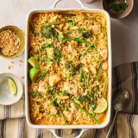 An oven dish with chicken noodles and vegetables on a table, ready to serve with a garnish of peanuts and lime on the side.