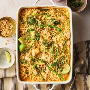 An oven dish with chicken noodles and vegetables on a table, ready to serve with a garnish of peanuts and lime on the side.