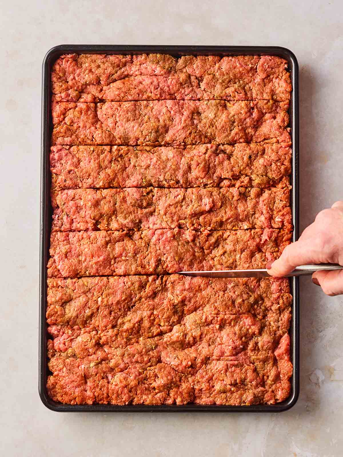 An oven tray with pressed beef mince on and a hand scoring lines into the slab with a knife.