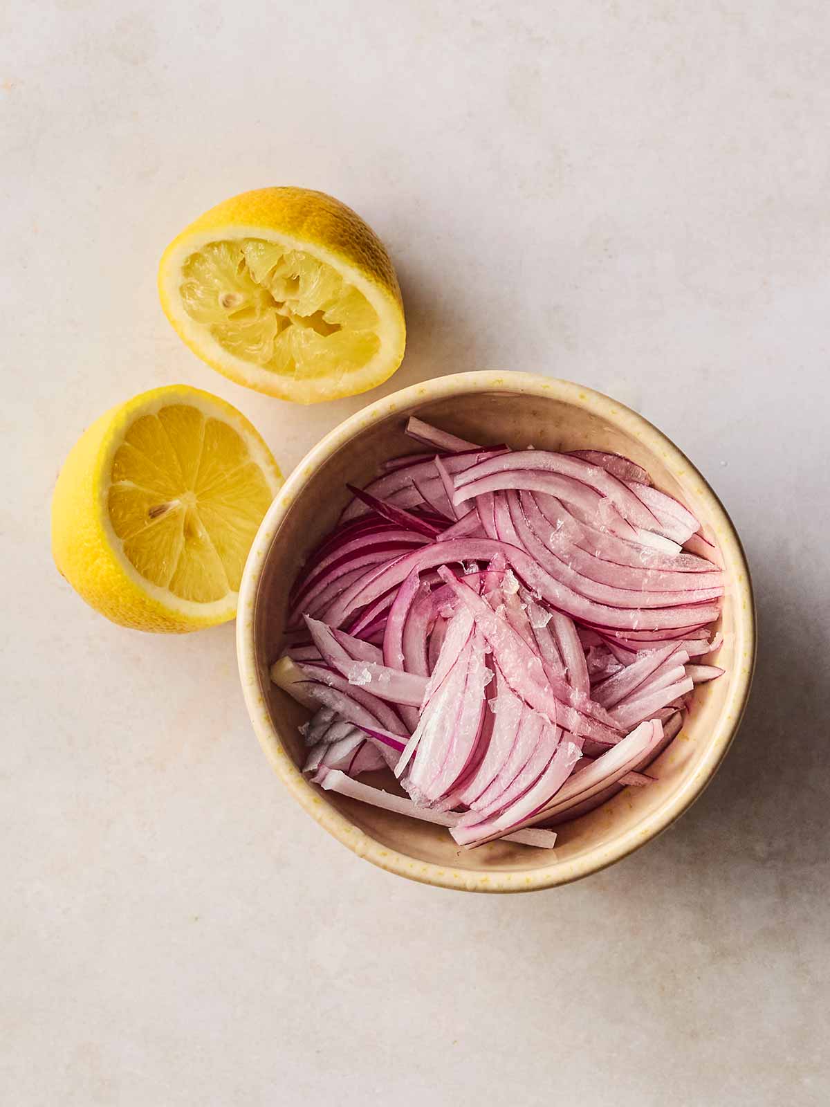 A sliced red onion in a dish with salt over and an empty half a lemon on its side next to it, showing it has been squeezed into the onion dish.