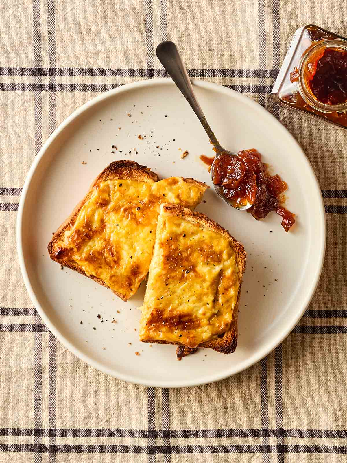 A plate with a slice of Welsh rarebit with a spoonful of chutney to the side, on a checked tablecloth.