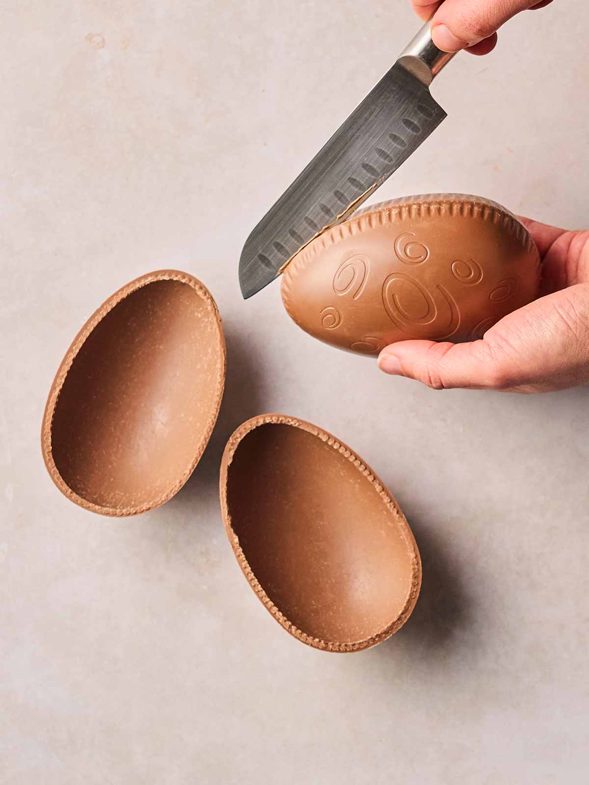 Two hands with a sharp knife slicing open a chocolate Easter egg, with a sliced one on a white surface next to them.