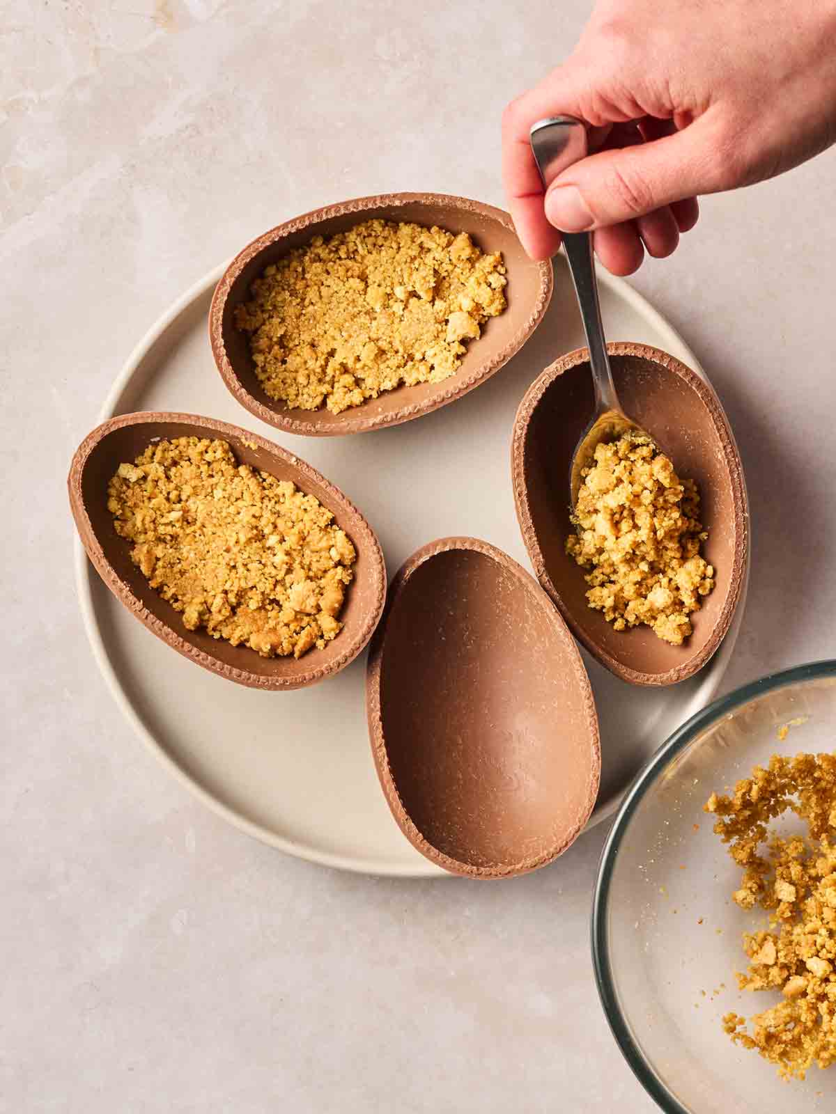Halved Easter eggs on a place, with a hand spooning in crushed biscuits to make a cheesecake base inside them.