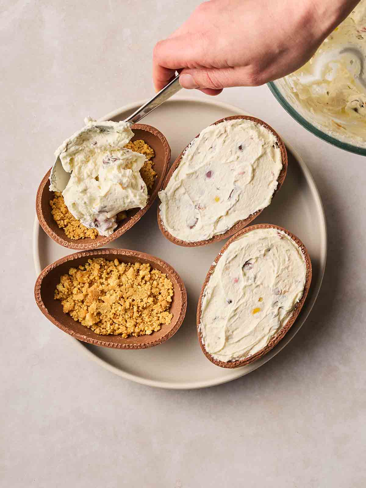 Four halved Easter eggs on a place, with a hand spooning in a creamy filling, with one showing a biscuit base for Easter Egg Cheesecakes.