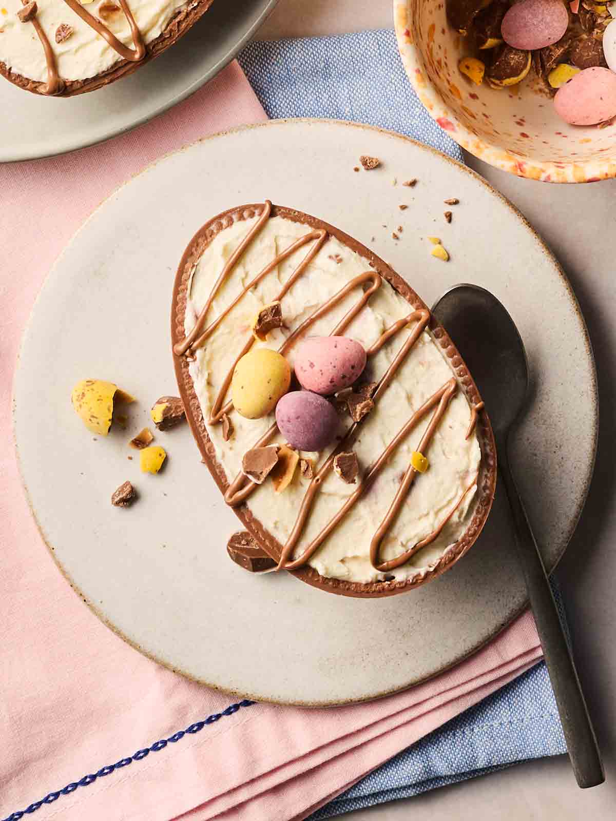 A cheesecake inside of an Eater Egg half, with mini eggs on top and swirls of melted chocolate. Ready to eat on a plate with a spoon.