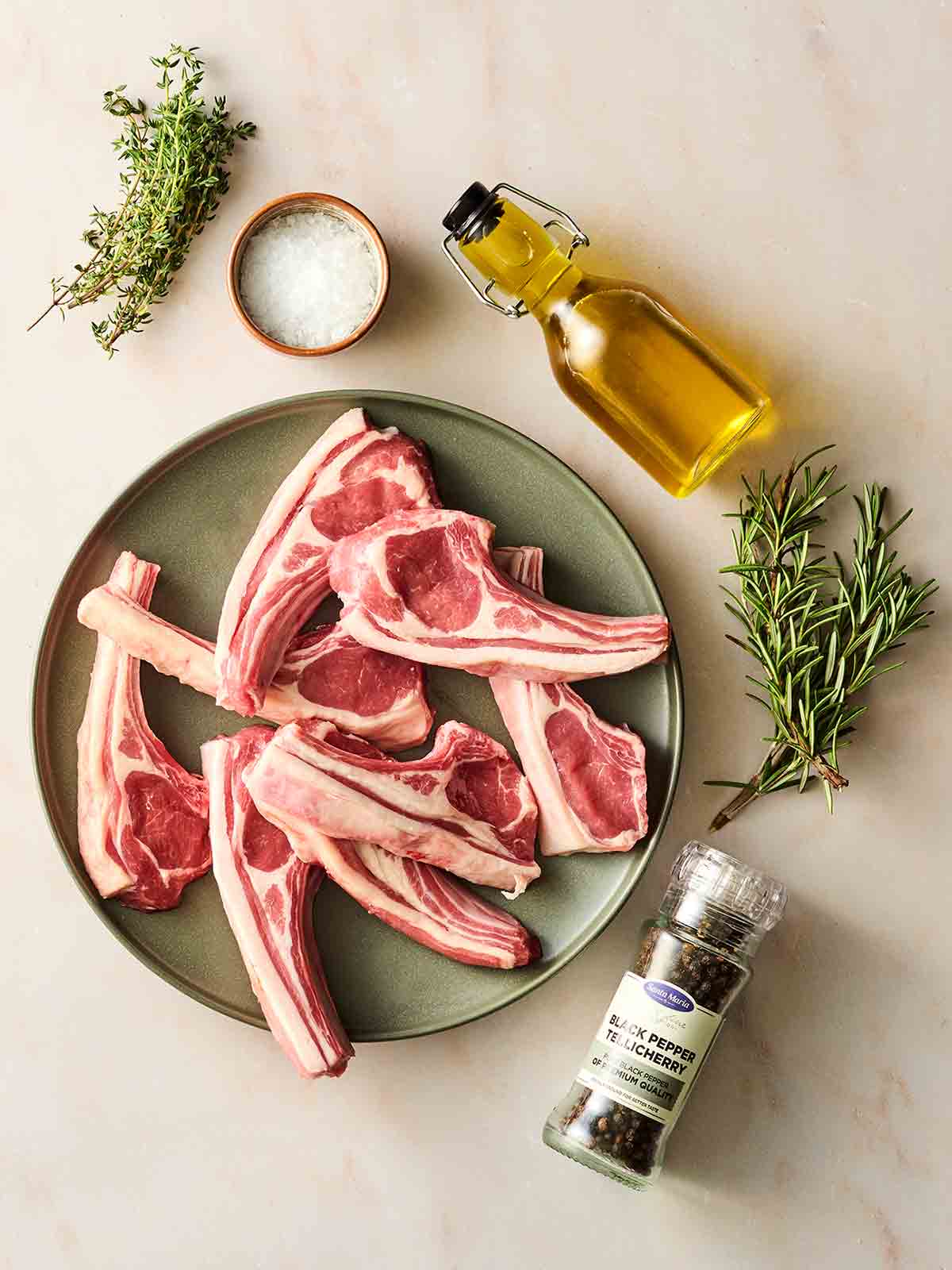 The ingredients for making perfect Lamb Chops on a white surface, including lamb, salt, rosemary, oil, thyme and black pepper.