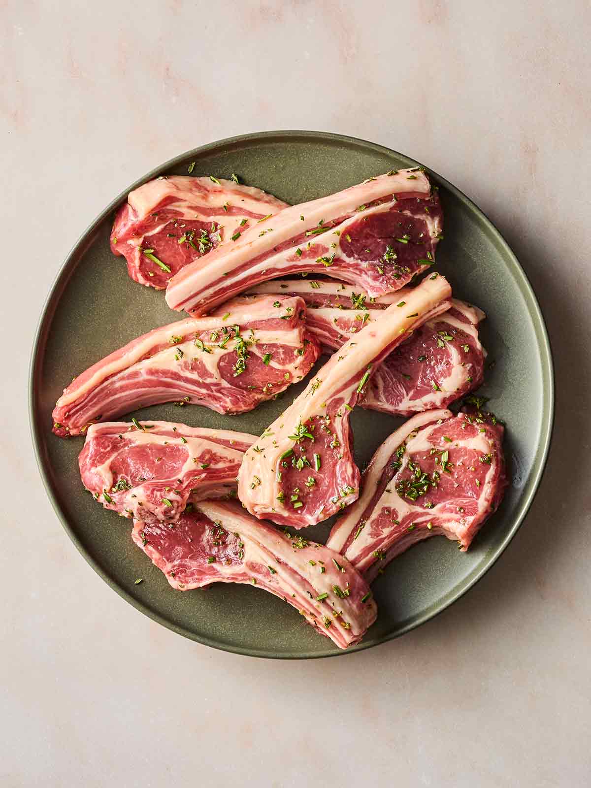 Raw lamb chops on a plate, with a garlic and oil herb marinade on top, ready to be cooked.