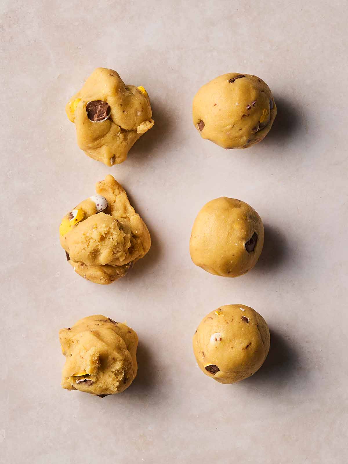 Six round balls of cookie dough with mini eggs on a white surface, ready to be flattened.