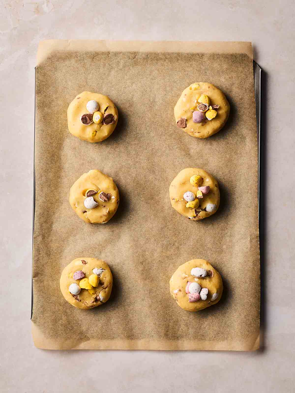 An oven tray lined with baking paper, with six round cookies with mini eggs, ready to be baked.