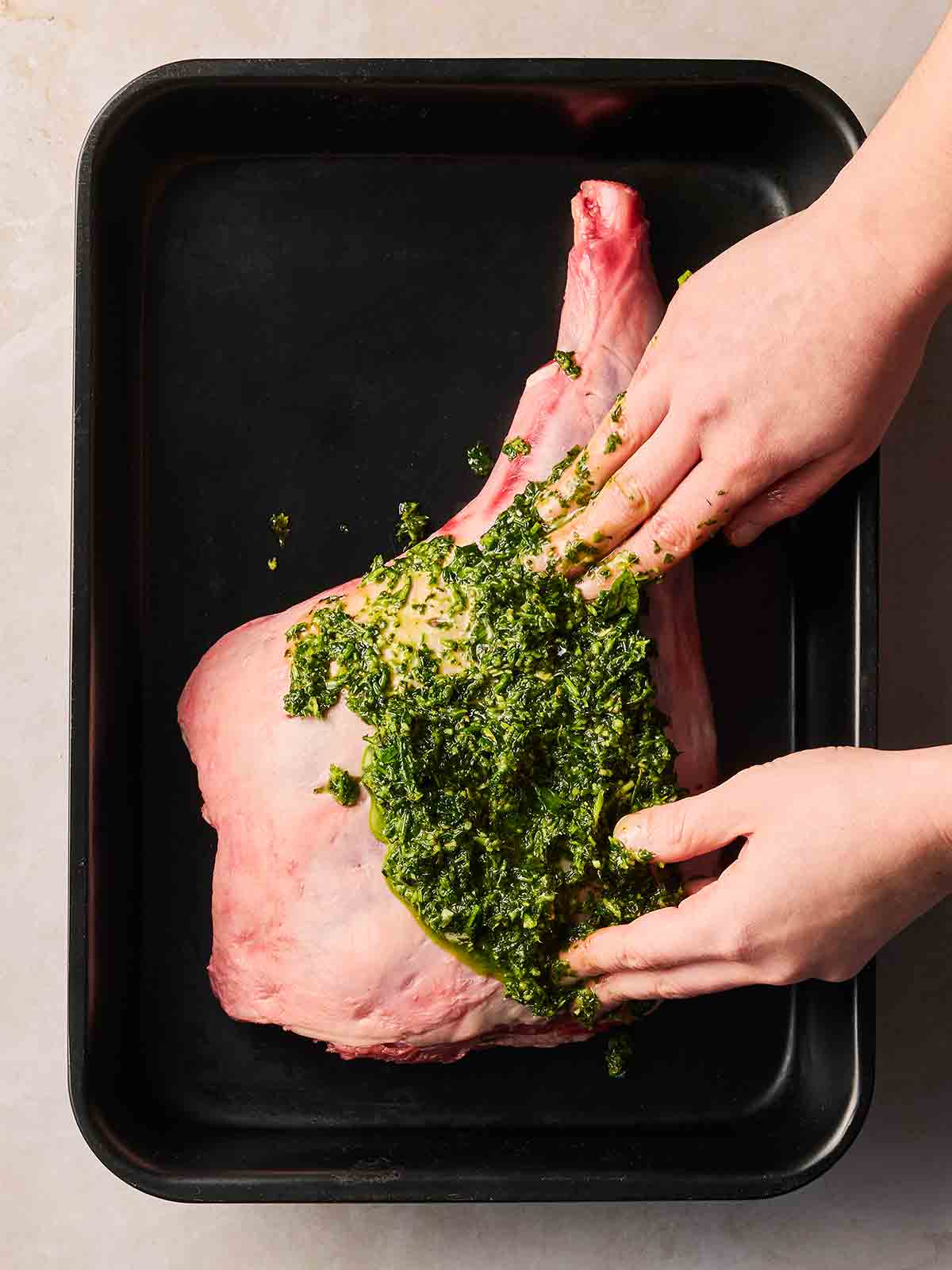 Two hands rubbing a green herby oil onto a raw leg of lamb that's sitting in a black oven tray.