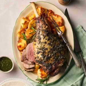 A roasted leg of lamb on a platter with roast potatoes to the side, with a few slices ready to eat, mint sauce to the side, and a carving knife and fork ready to slice up the rest.
