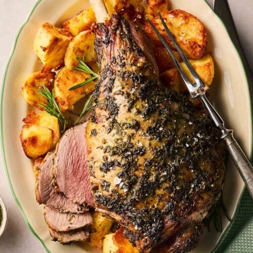 A roasted lamb leg with a green herb rub over the top, ready to carve on a platter with roast potatoes.