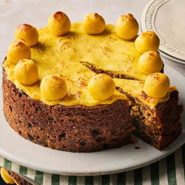 A close up of a traditional Simnel Cake, topped with marzipan, on a white plate, with a slice cut out.