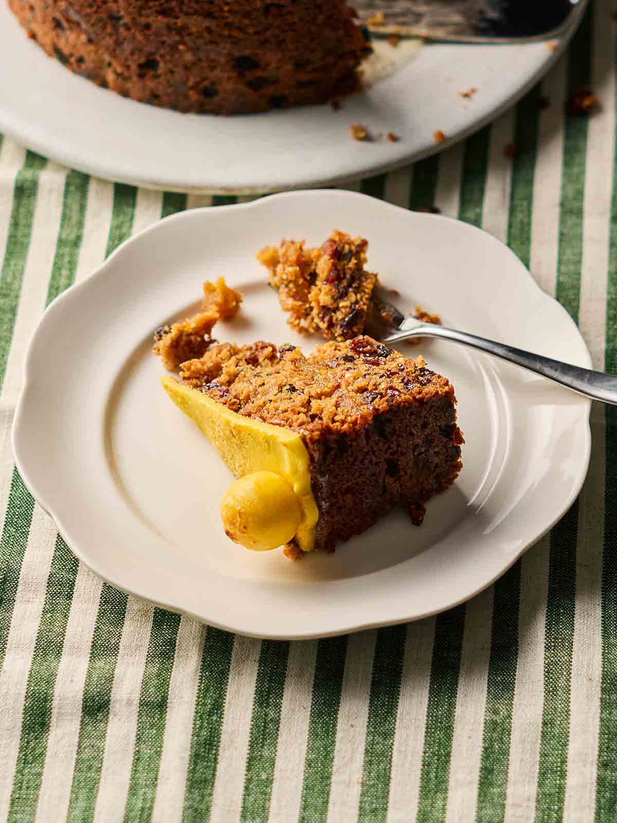 A slice of fruity Simnel Cake on a white cake plate with a fork stuck in.