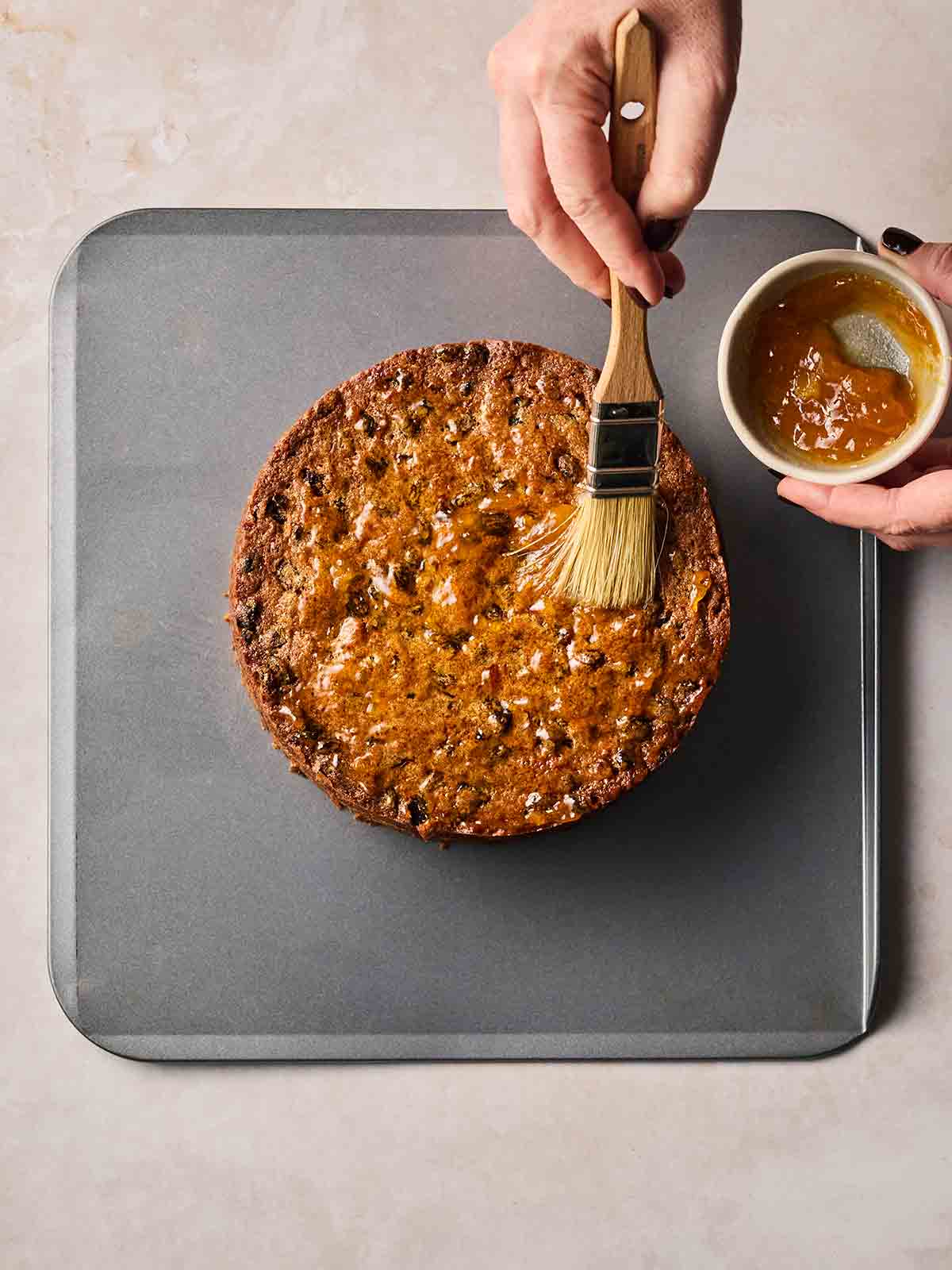 A baked fruit cake on an oven tray, with two hands brushing over apricot jam.