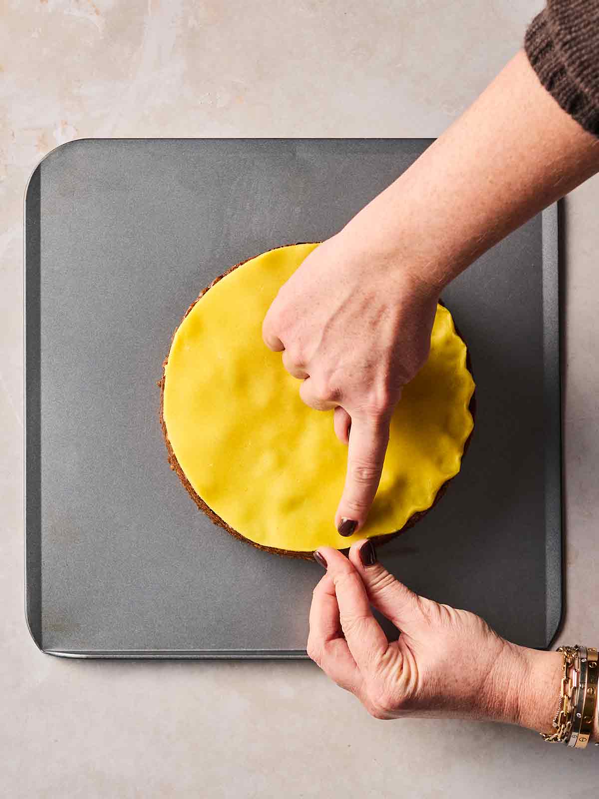 A bird's eye view of a Simnel Cake, decorated with marzipan topping, with two hands pinching it in at the edges.