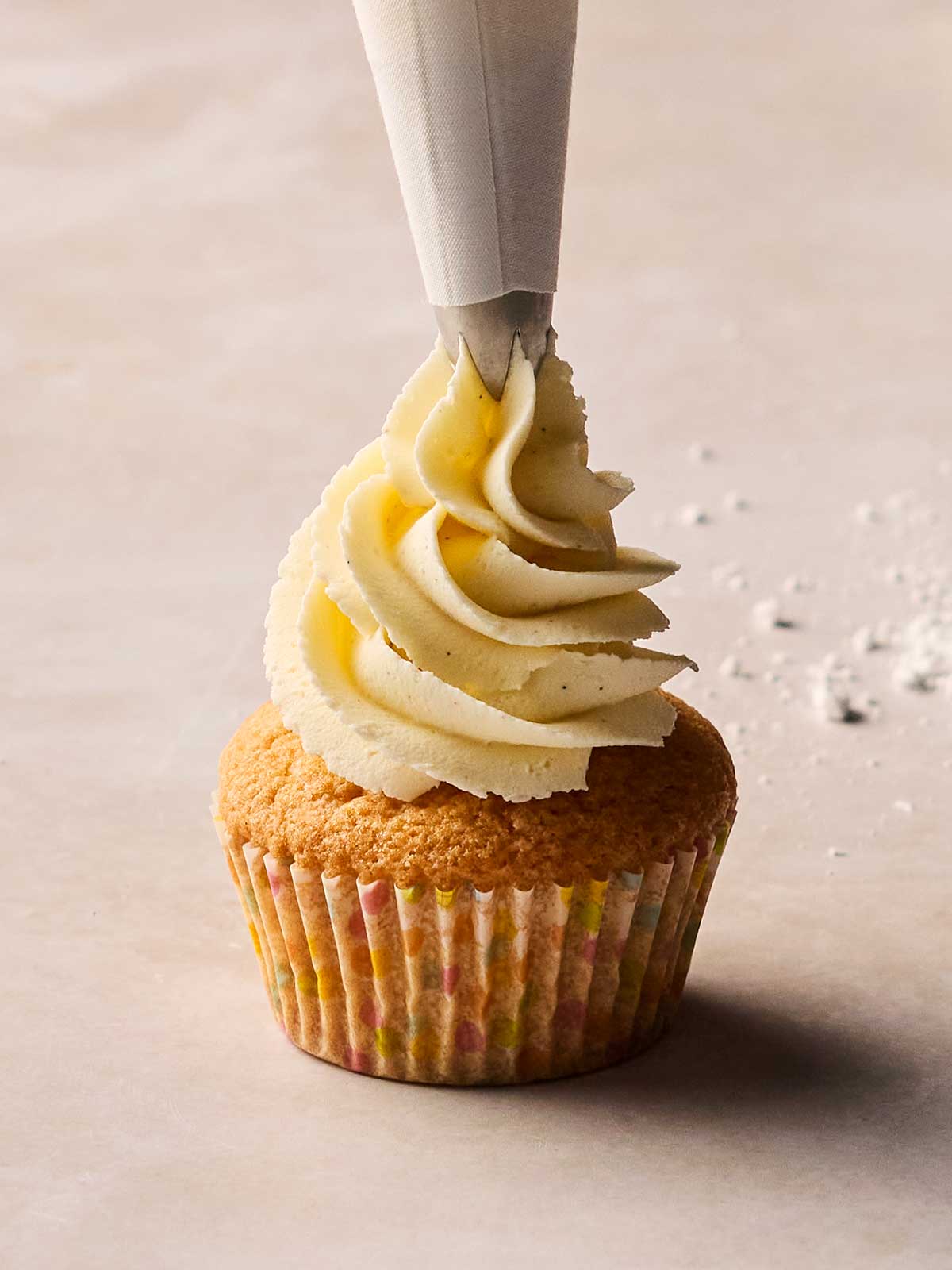 A cupcake being iced with buttercream frosting through a nozzle.