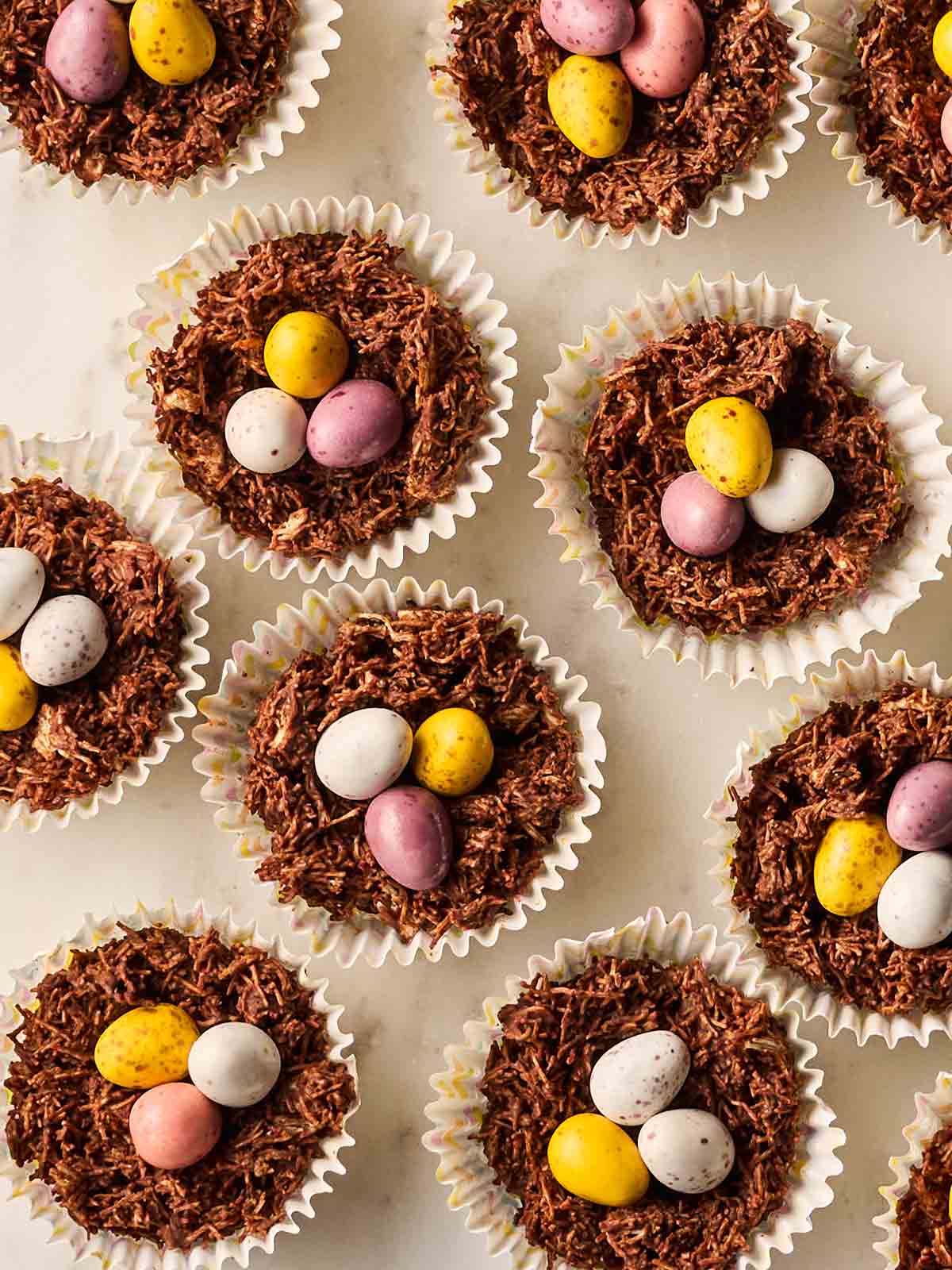 Rows of Easter Nests with chocolate and mini eggs in cupcake cases, on a white surface.