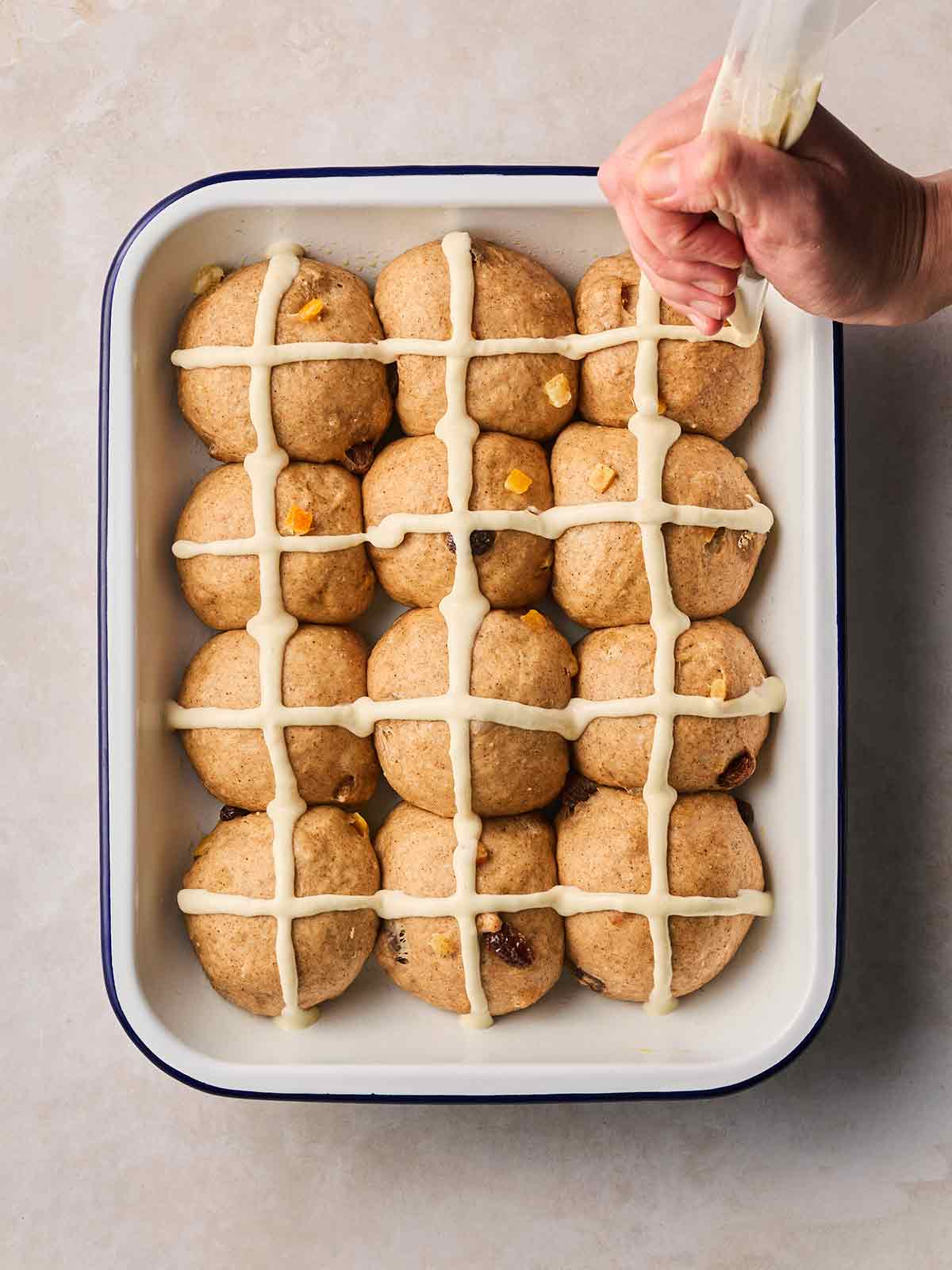 A hand piping crosses onto unbaked Hot Cross Buns in a white oven dish, on a white surface.