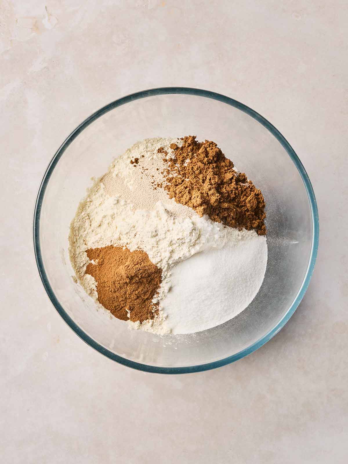 A glass bowl with flour, sugar, yeast and spices inside, ready to mix.