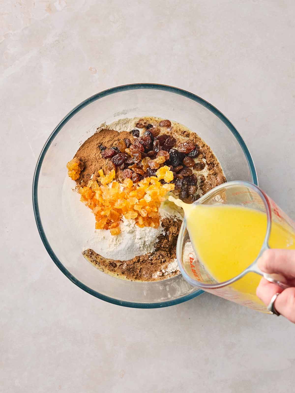 A hand pouring melted butter and milk from a jug into a glass bowl with flour, sugar, spices and mixed dried fruit in.