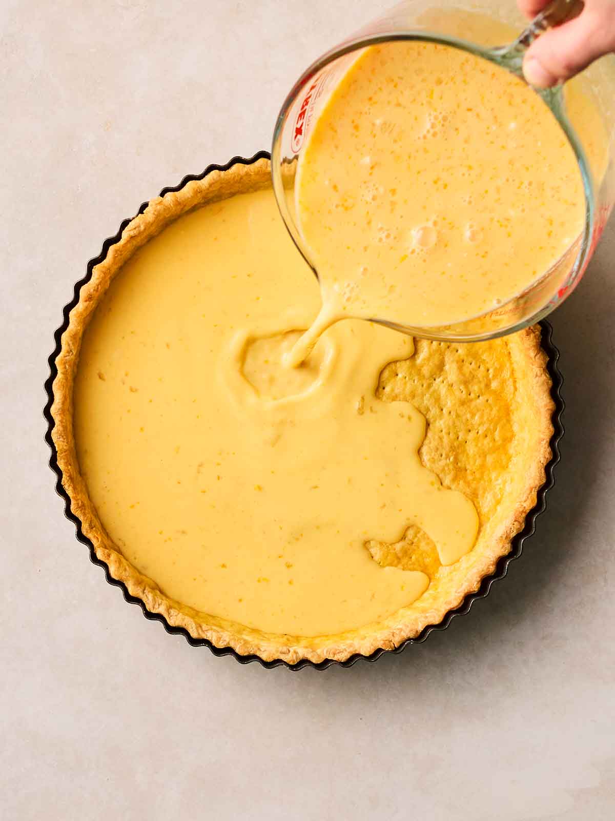A hand pouring a jug of yellow lemony mixture into a shortcrust pastry pie tart shell to make a Lemon Tart.