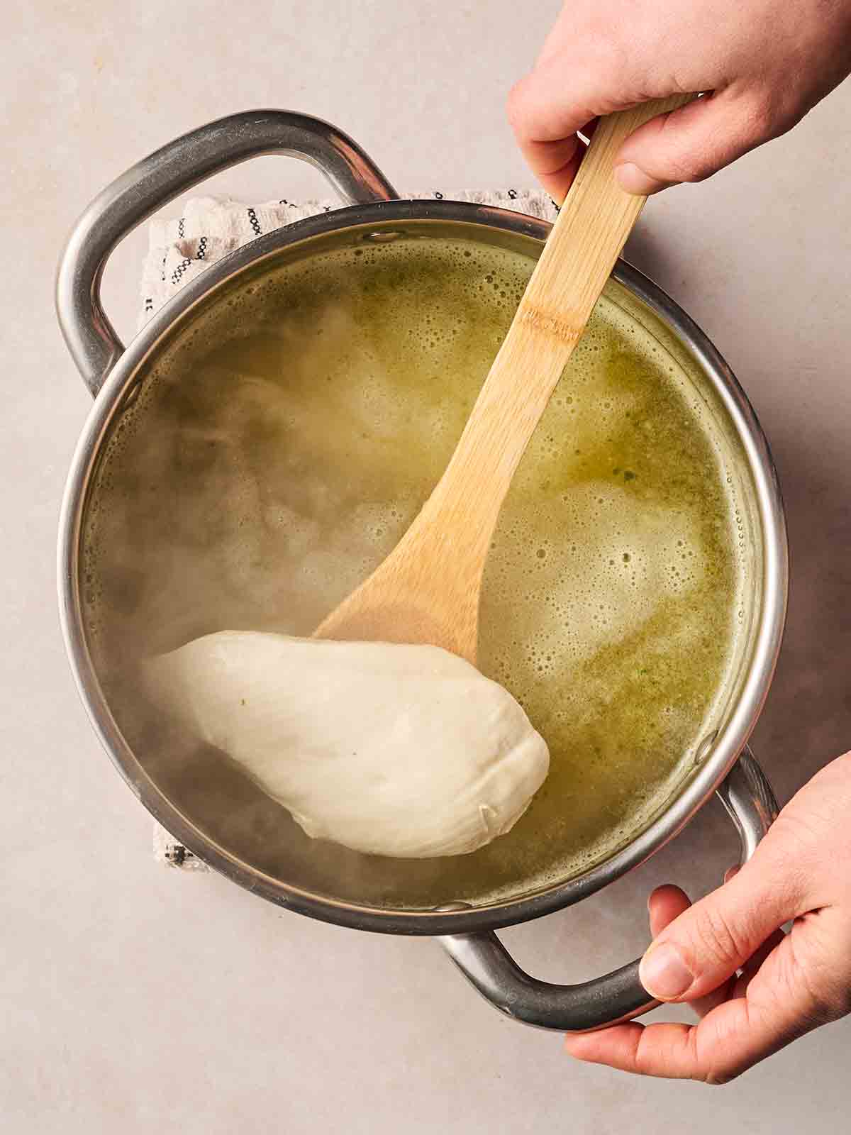 A pan filled with stock, with a hand spooning a cooked chicken breast out of the liquid, and the other hand holding the pan handle.