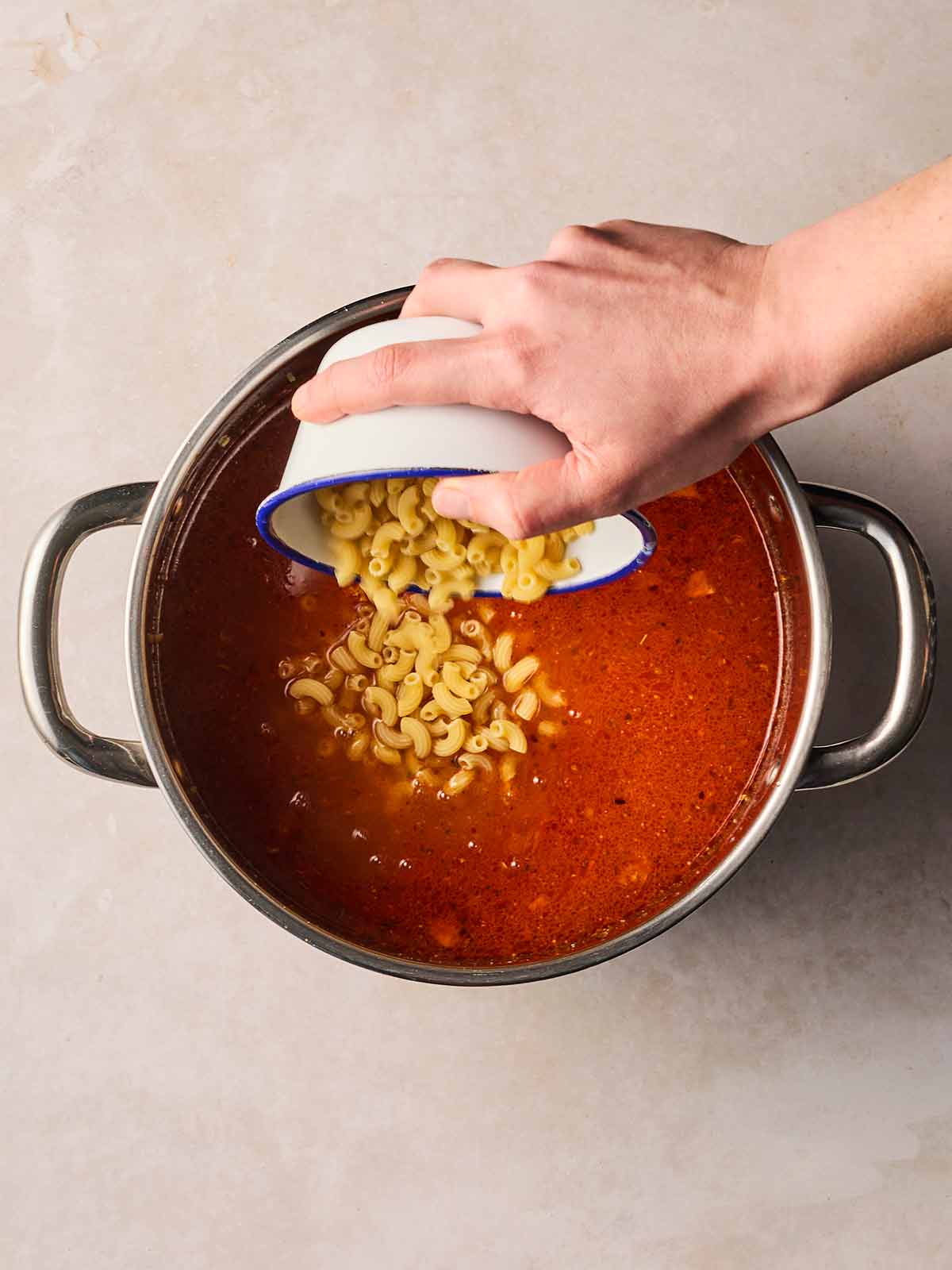 A hand pouring in macaroni pasta into a bowl of tomatoey soup.