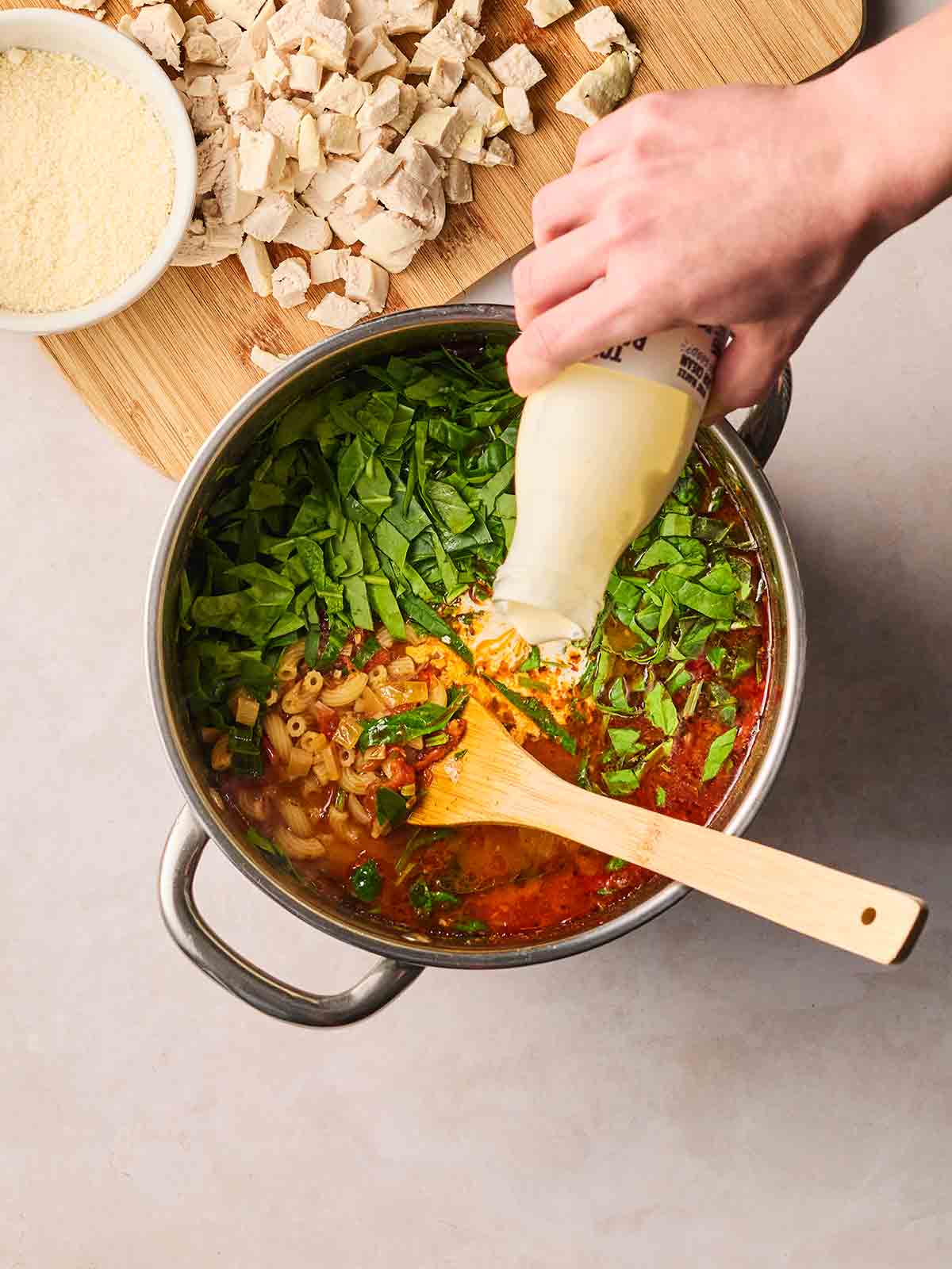 A hand pouring cream into a pan of Marry Me Chicken Soup, with a board of diced cooked chicken to the side, ready for it to go in.