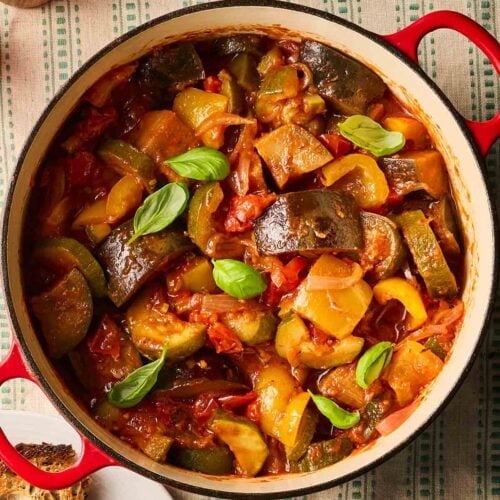 A Mediterranean Vegetable Stew known as Ratatouille in a large pan, sitting on a tablecloth with green stripes, and bread and basil to the side.