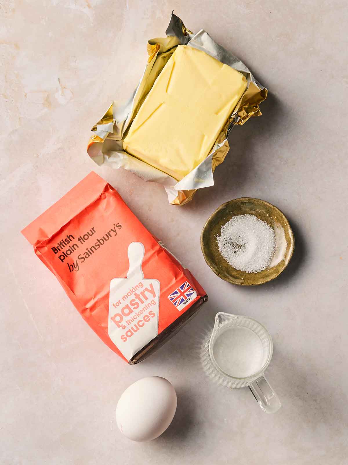 The ingredients for making shortcrust pastry on a white surface, including plain flour, butter, an egg, water and some salt.