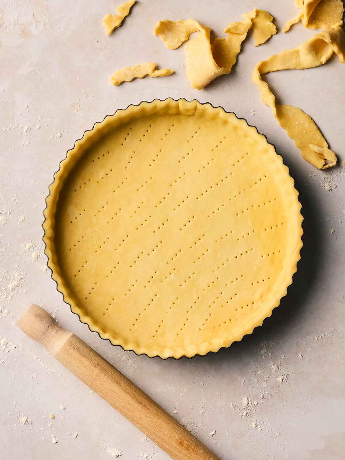 A flan dish with unbaked homemade shortcrust pastry inside with a rolling pin to the side and offcuts of pastry, on a white surface.