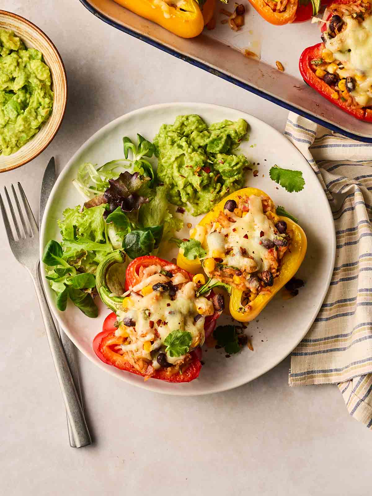 A plate with stuffed peppers and cheese, with salad and guacamole to the side on a white plate, with cutlery to the side.