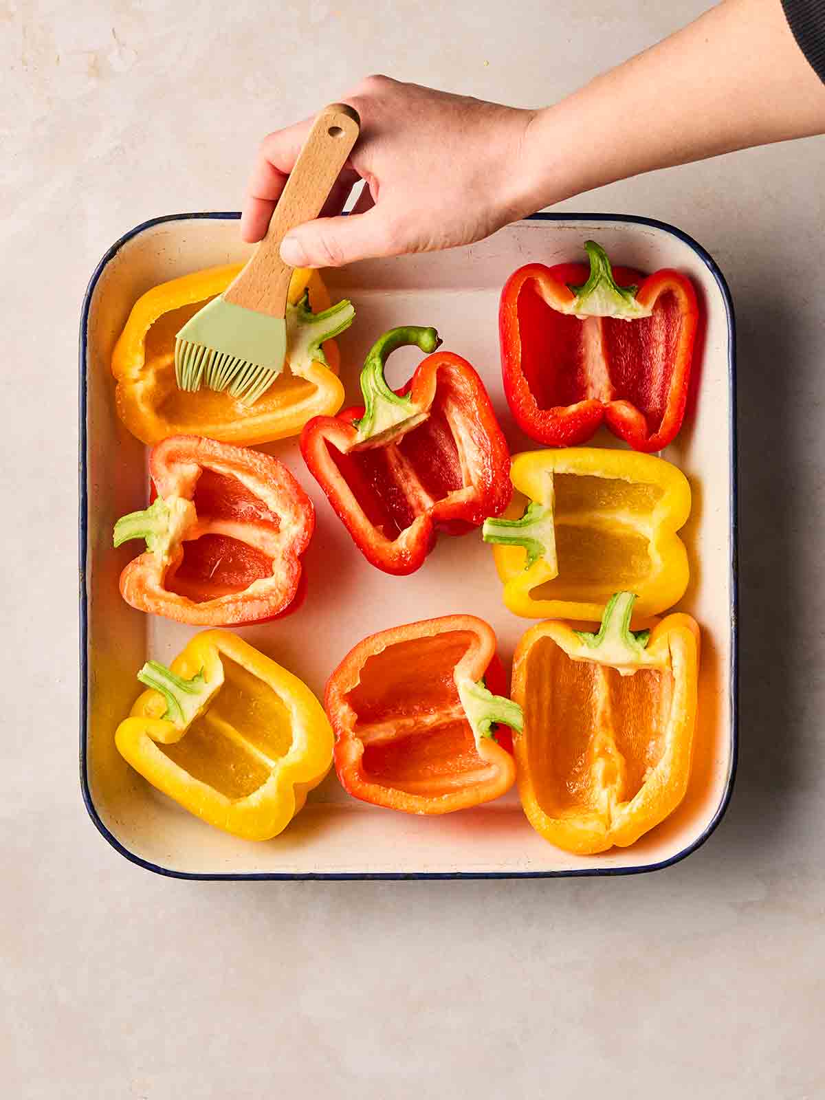 A hand brushing halved peppers in an oven dish with oil.