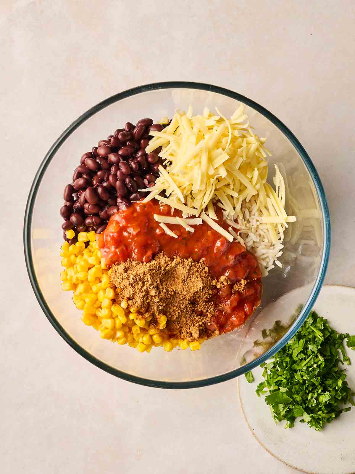 A glass bowl with ingredients inside, ready to be mixed. Including cheese, salsa, beans, sweetcorn and spices.