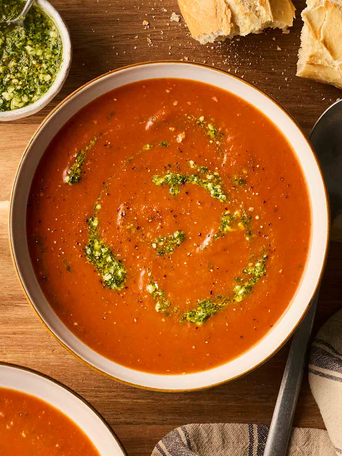 A bowl of homemade tomato soup on a table, with pesto on top, a spoon to the side and bread to dip in.