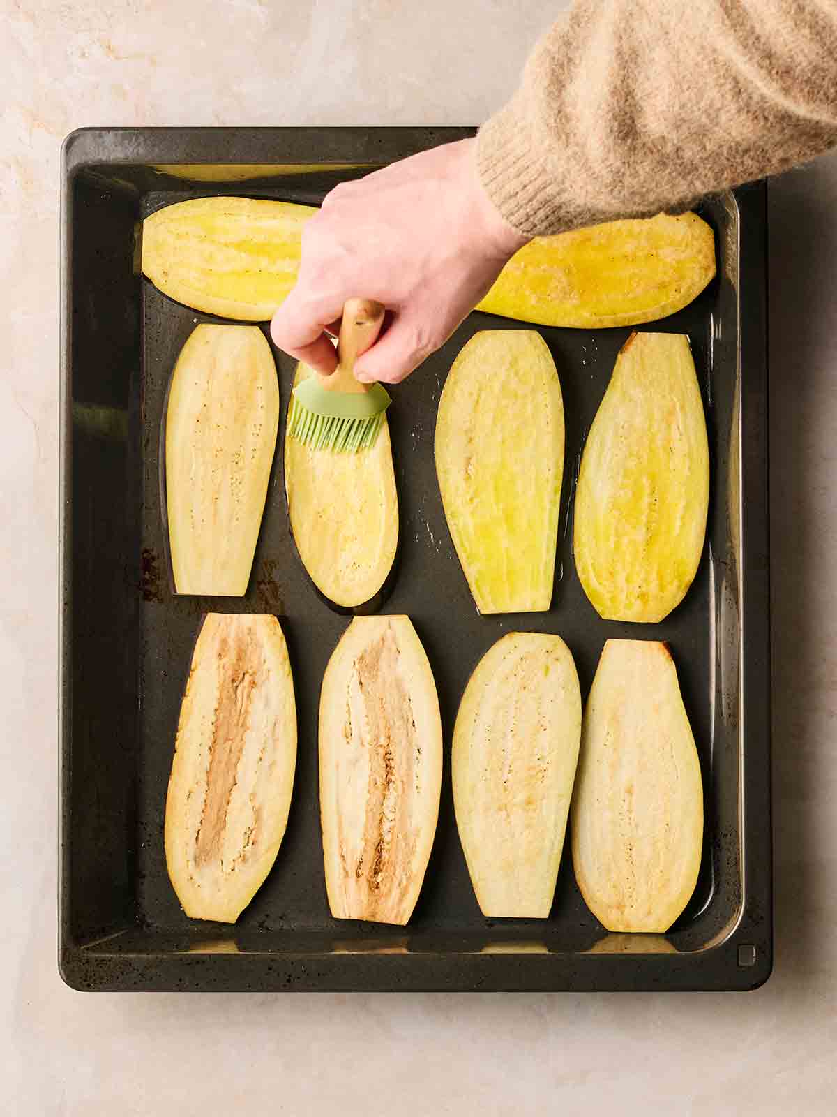 A hand brushing oil over sliced aubergines on an oven tray.