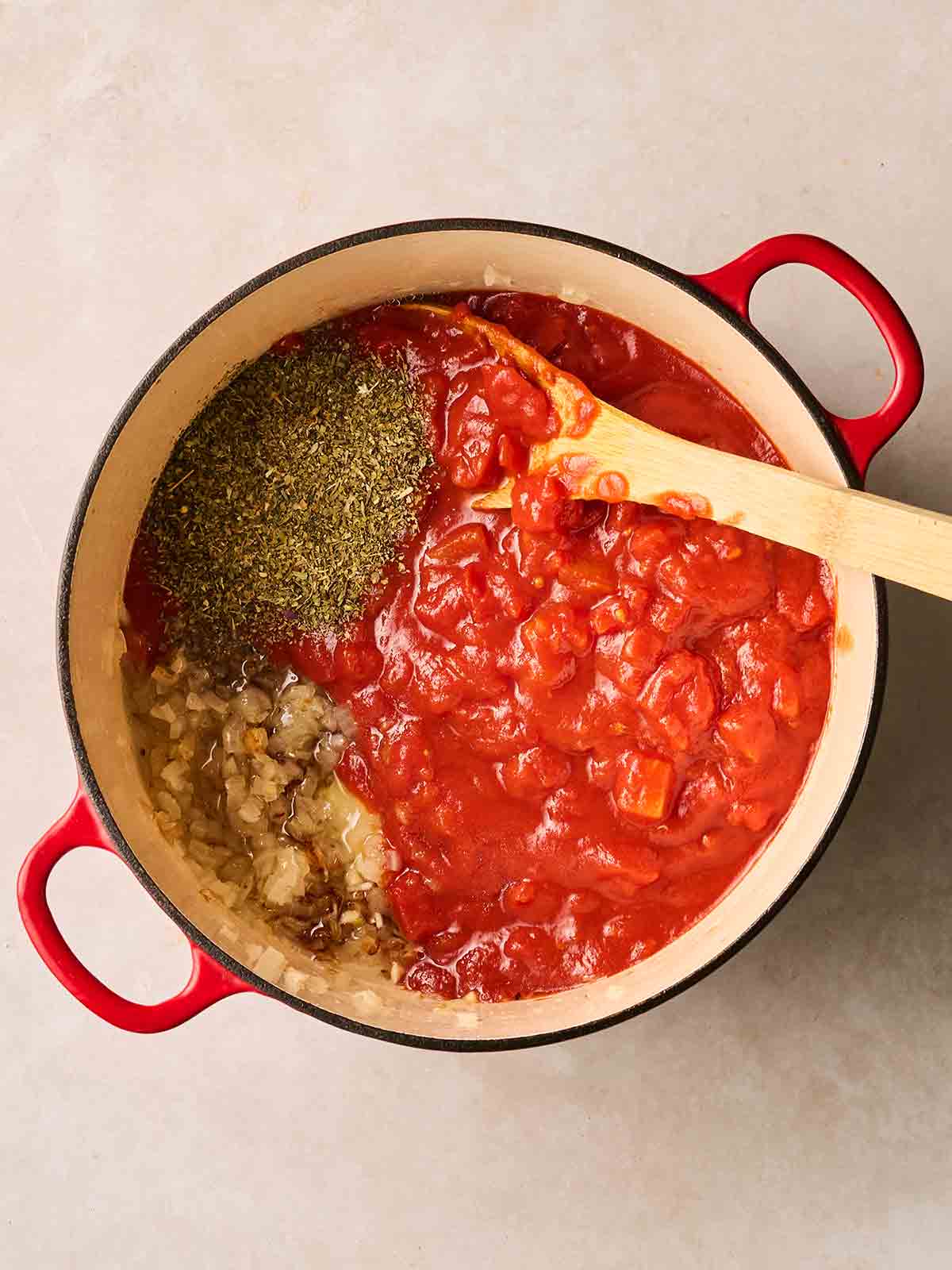 A large pan of tomato sauce with herbs and onions being stirred together.