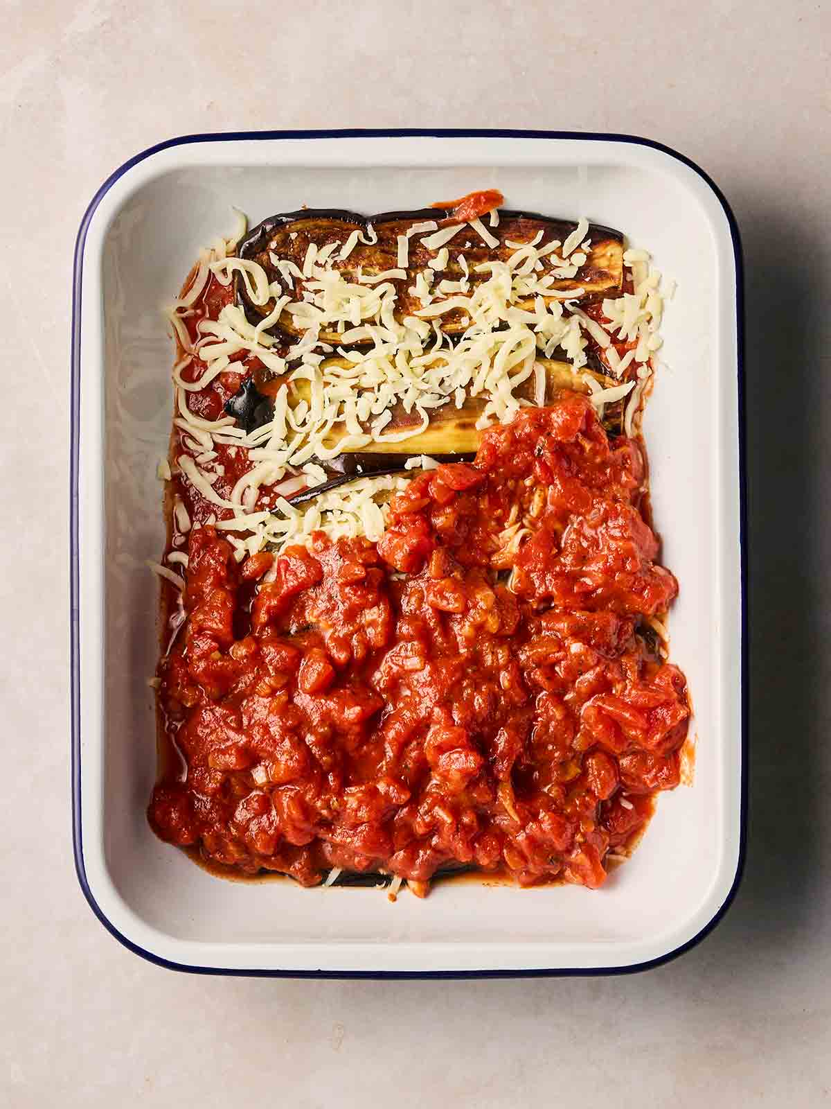 A white oven dish showing ingredients layered up for Eggplant parmesan, with tomato sauce, aubergine slices and mozzarella cheese.