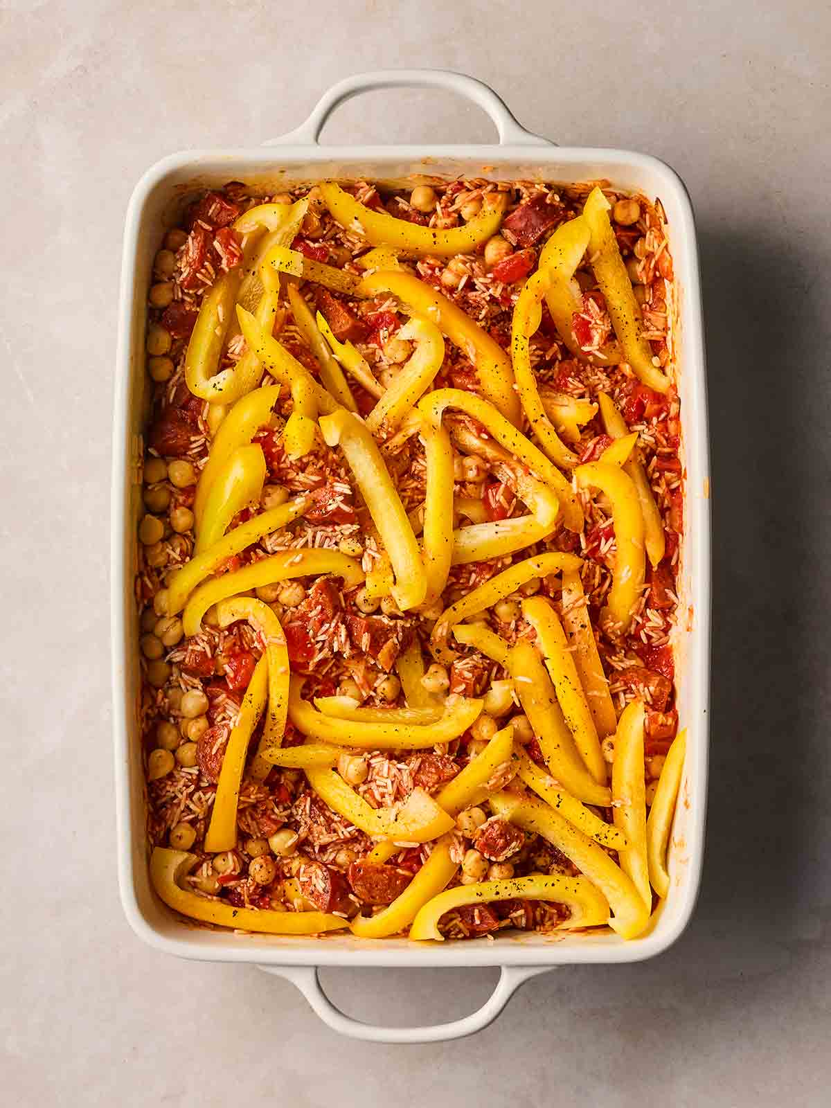 An oven dish with ingredients in, ready to bake together, including rice, chorizo, onion, chickpeas and sliced yellow peppers.