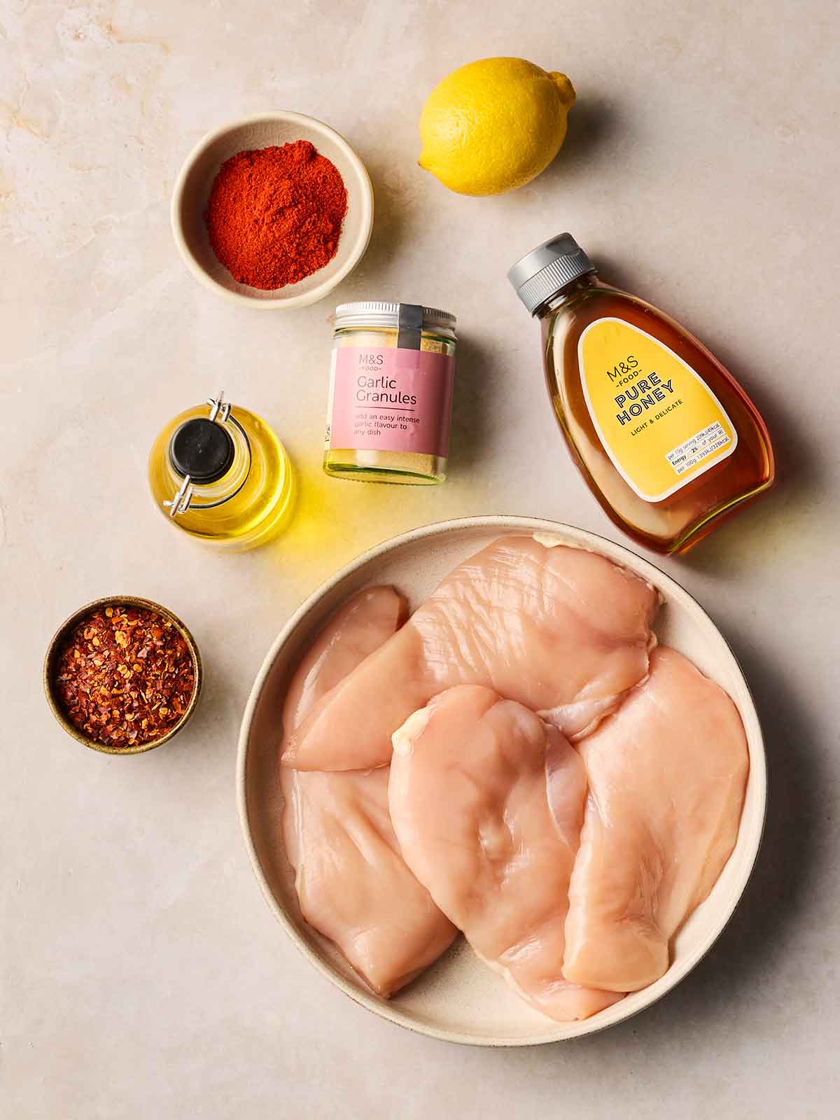 The ingredients for making a chicken dish with a marinade on a white surface, including raw breasts, chilli flakes, paprika, honey, a lemon, garlic granules and oil.