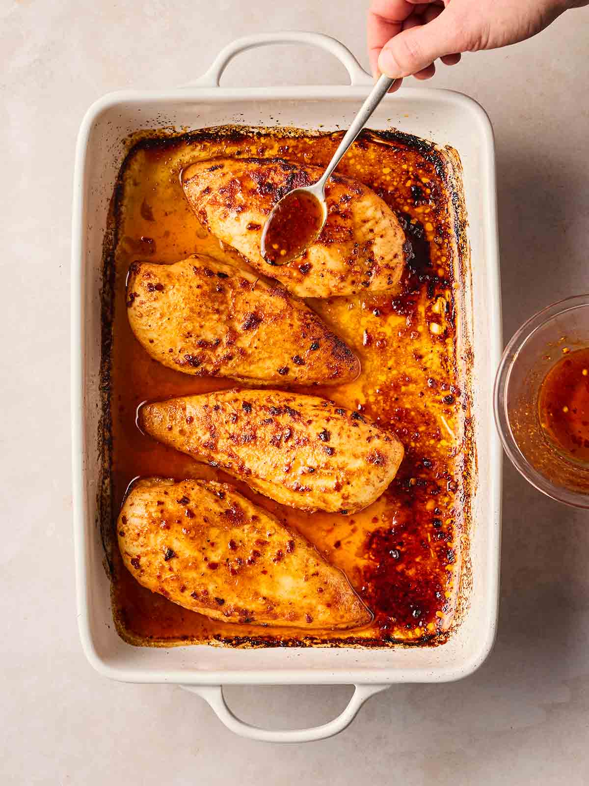 A hand sprinkling a glaze over chicken breasts with a spoon for a baked Divorce Me Chicken dish.