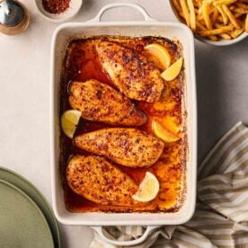 An oven dish with four baked chicken breasts on table, with a bowl of chips to the side, ready to serve.