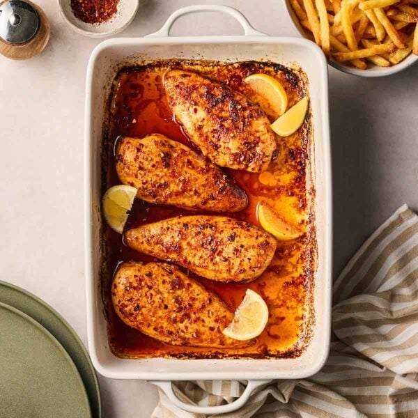 An oven dish with four baked chicken breasts on table, with a bowl of chips to the side, ready to serve.