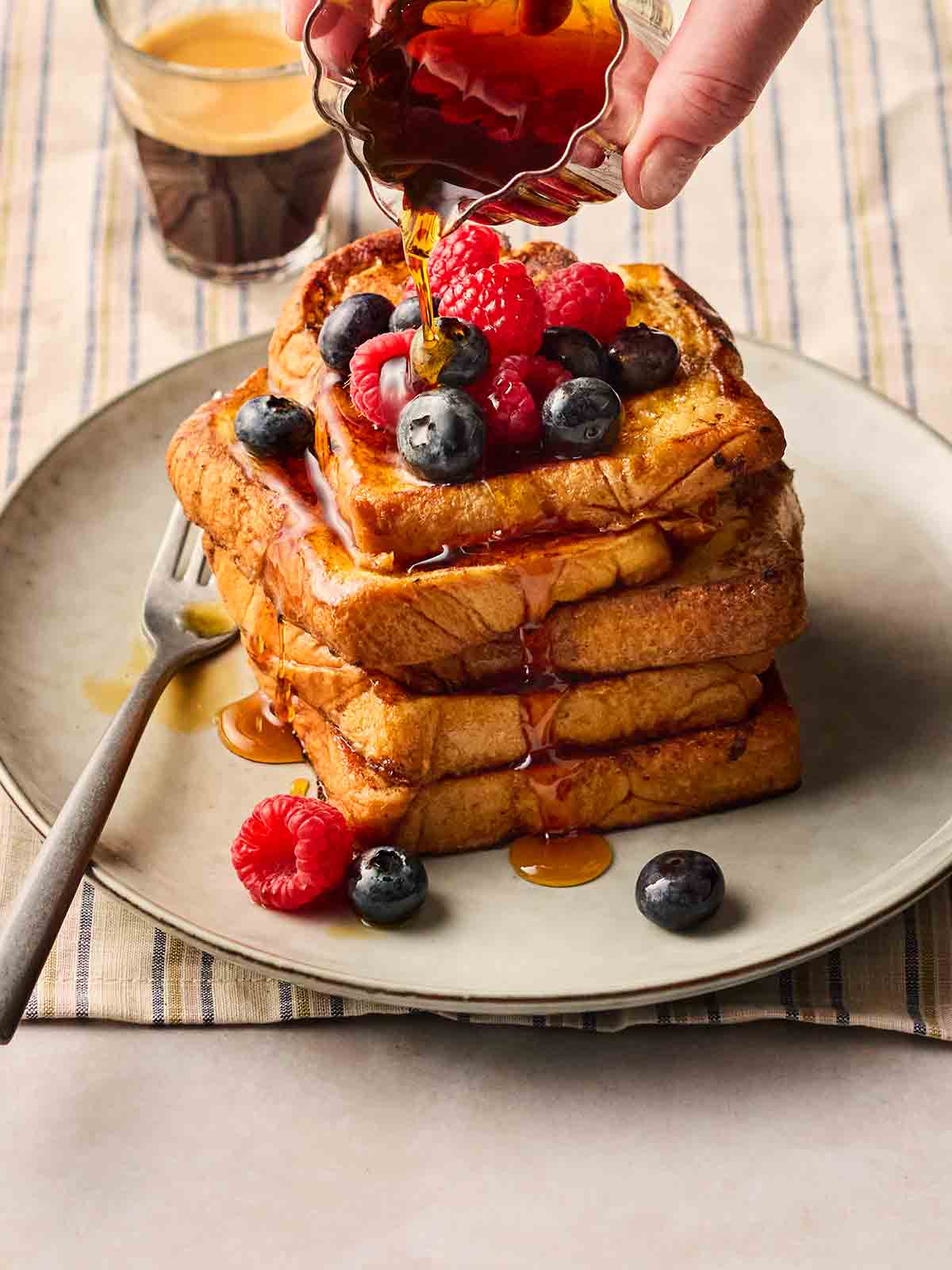 A stack of French Toast on a plate, topped with berries and a hand pouring maple syrup over the top.