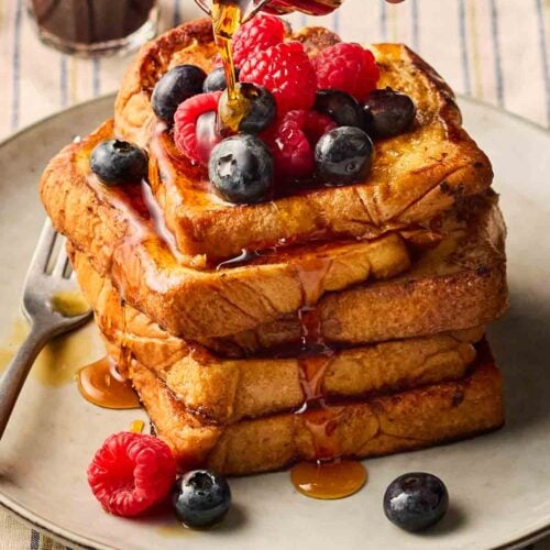 French toast stacked on a plate on a set table, with berries on top and maple syrup being poured over.