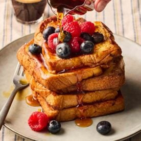 A close up and side angle of a stack of French toast with berries over and maple syrup being poured on.